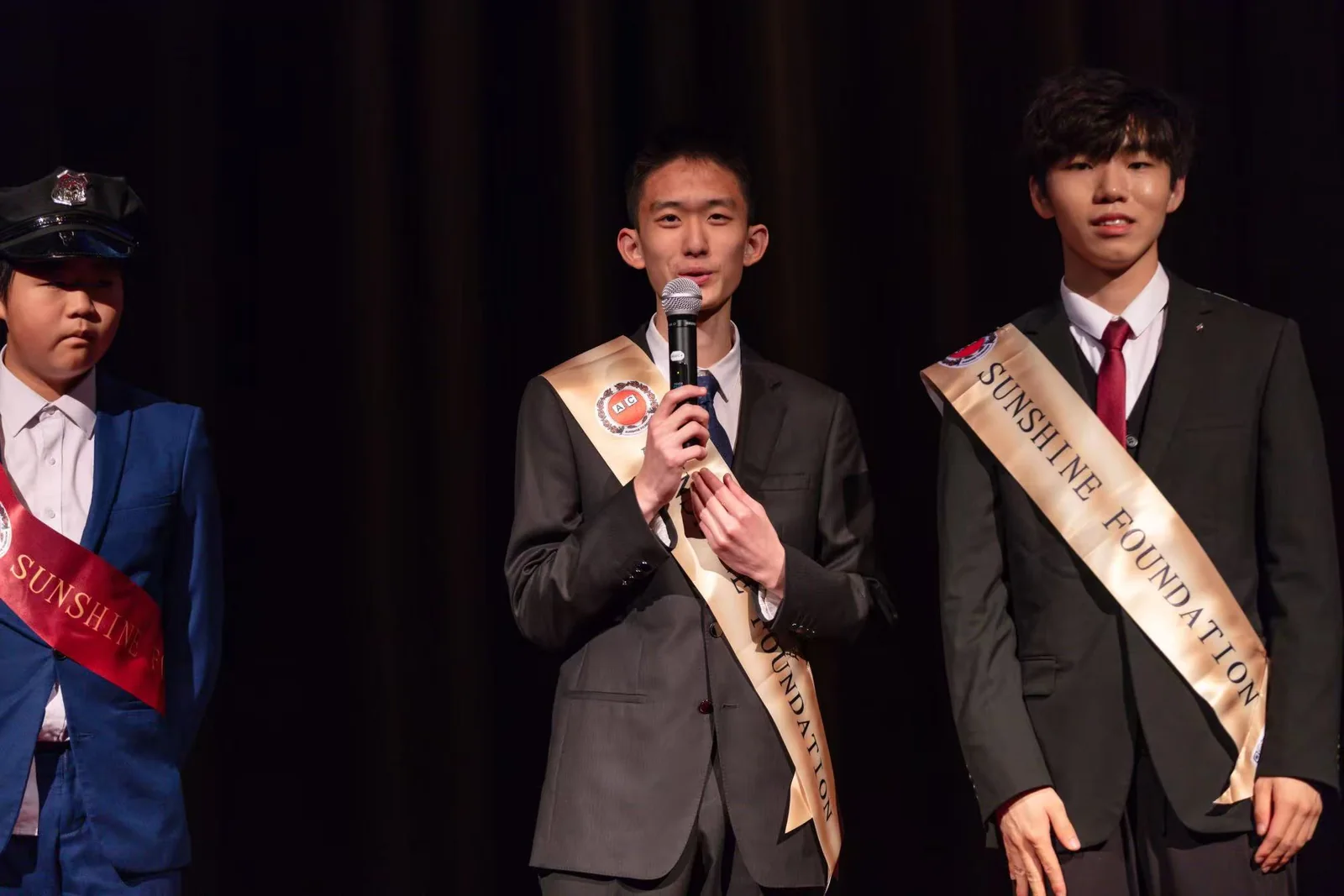 Terry speaking into a microphone on a darkened stage, wearing a Sunshine Foundation sash across his suit, with another board member beside him.